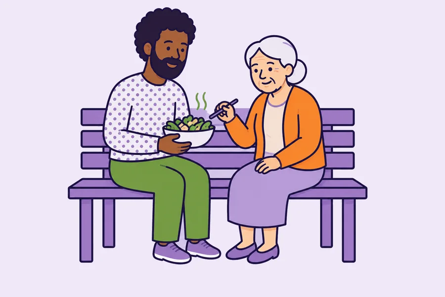 A support worker and an older woman sitting together on a bench, sharing a meal she is eating herself.